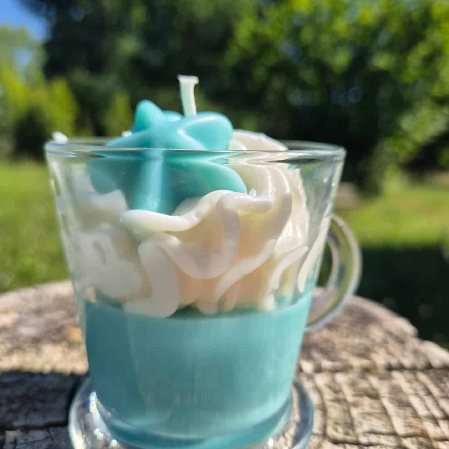Bougie parfumée artisanale au Monoï dans une tasse en verre transparente, décorée d’une chantilly blanche et de détails bleu lagon, posée sur une souche en bois en plein soleil avec un fond de jardin verdoyant flou.