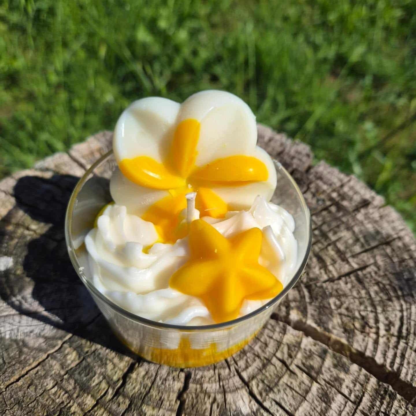 Bougie parfumée artisanale Monoï Vanillé avec chantilly blanche et fleurs décoratives jaunes et crème dans un contenant en verre, posée sur une souche en bois en extérieur.